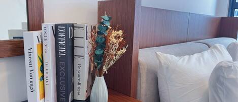 Una estantería ordenada con libros y un jarrón de flores secas, ubicada junto a una cama con ropa de cama blanca.