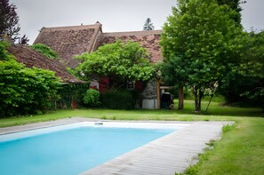 A swimming pool with a wooden deck, surrounded by greenery and a building with a tiled roof.