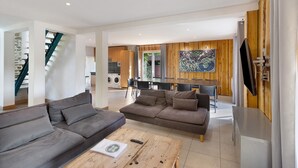 A modern living room with a sofa, coffee table, and a TV. There's a staircase, a dining area, and a kitchen in the background.