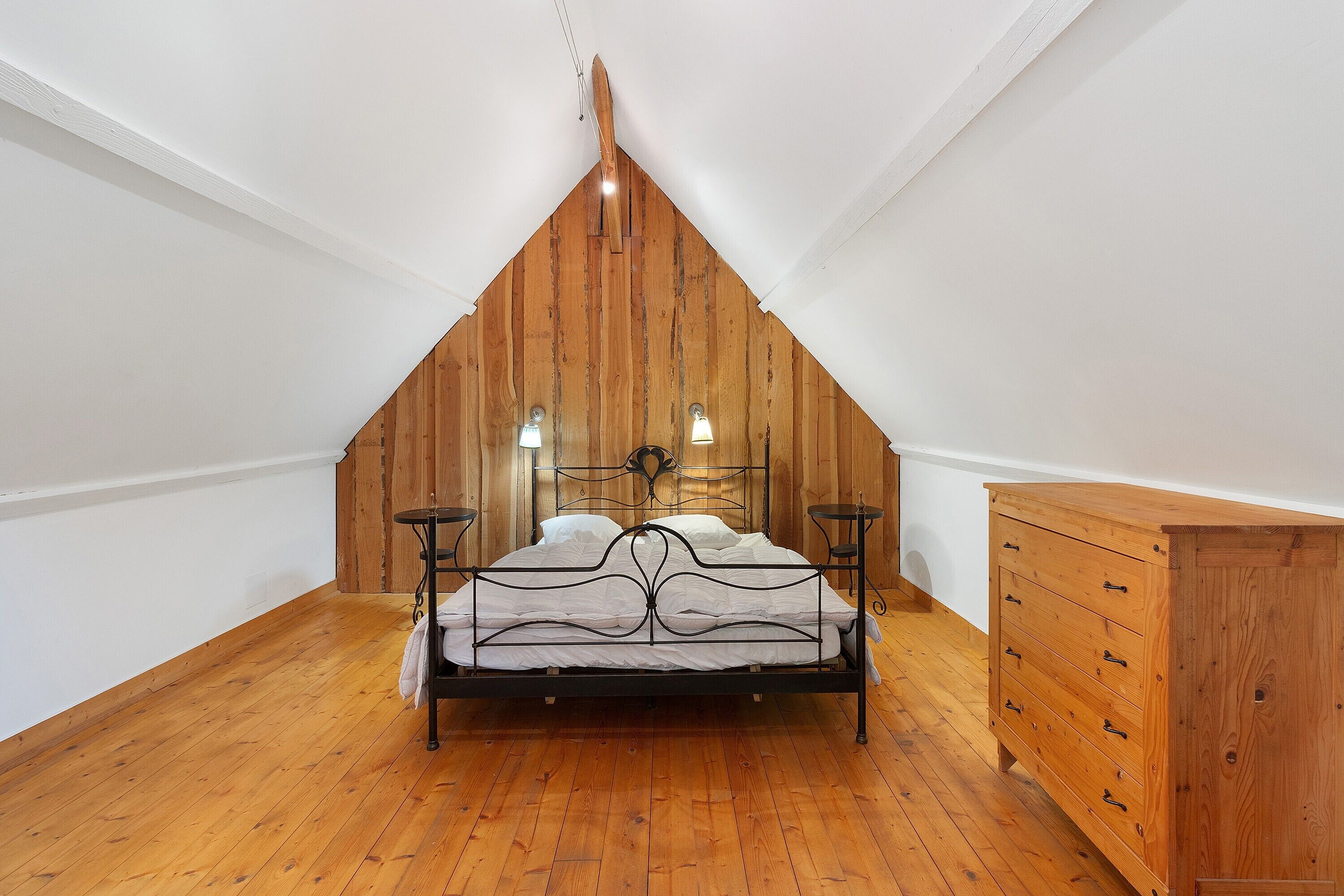 Ein Schlafzimmer mit schrägem Dach, einem Bett, Nachttischen und einer Holzkommode.