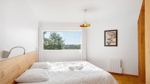 A bedroom with a wooden headboard, a bed with white bedding, a window with a view of greenery, a pendant light, and a radiator. - Modern holiday home with private pool and spa in the Périgord countryside (Angoisse)