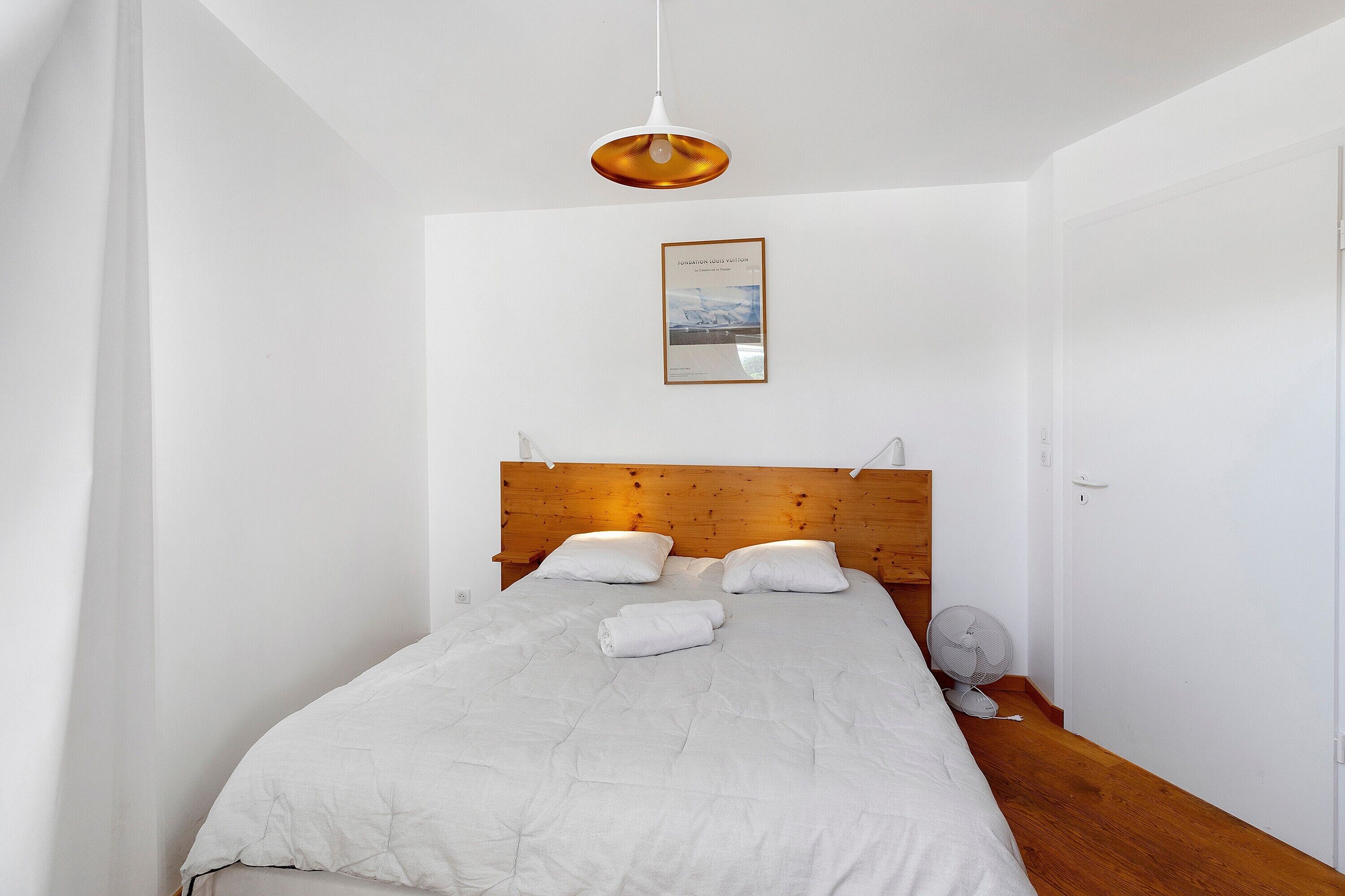 Een net opgemaakt bed met een houten hoofdbord, een nachtkastje, een wandlamp en een ingelijst kunstwerk aan de muur.