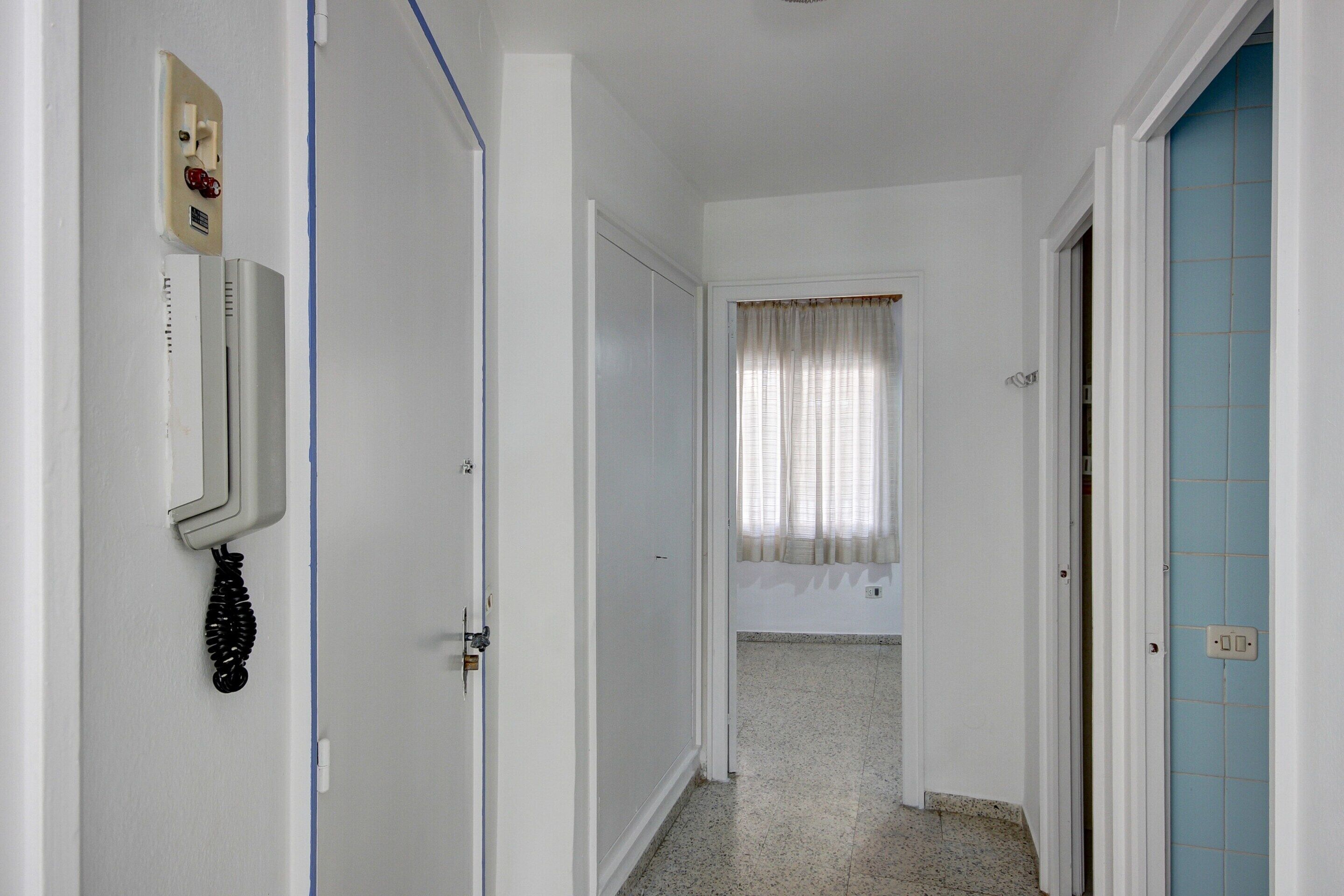 A narrow hallway with white walls, a door with a light switch, and a blue-tiled wall on the right.