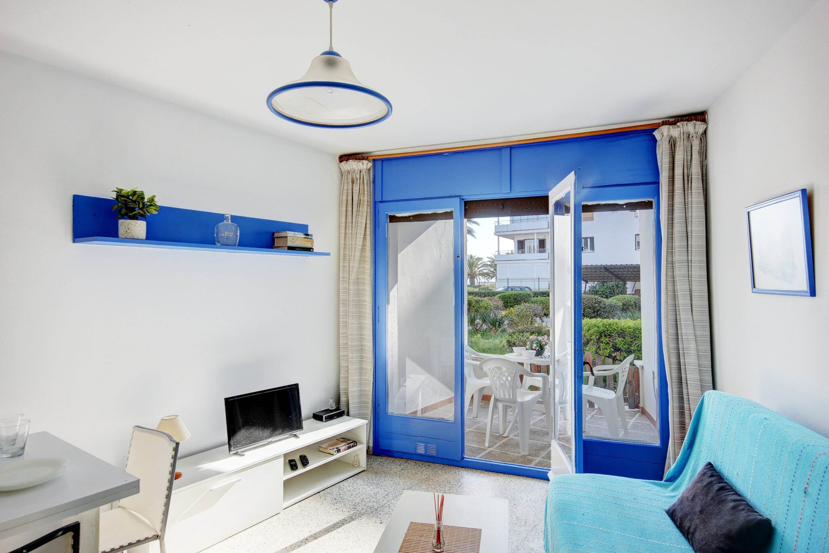 A room with a blue sofa, a TV on a white stand, a small table with a lamp, a ceiling light, and a sliding glass door leading to an outdoor area with chairs.