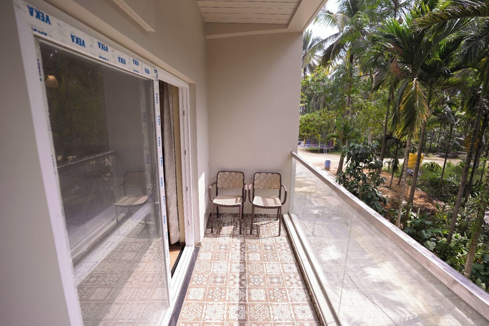 A balcony with two wicker chairs, a patterned tile floor, and a view of a tropical garden with palm trees.