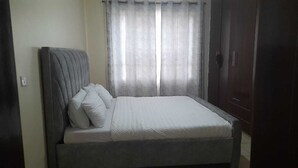 A bed with a grey headboard and white bedding, a window with sheer curtains, and a dark wooden wardrobe.