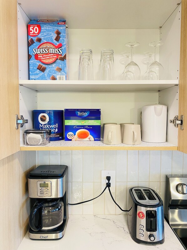 A kitchen with a coffee maker, toaster, and a shelf with a box of Swiss Miss chocolate milk, a box of Tetley Orange Pekoe tea, and a Maxwell House coffee jar.