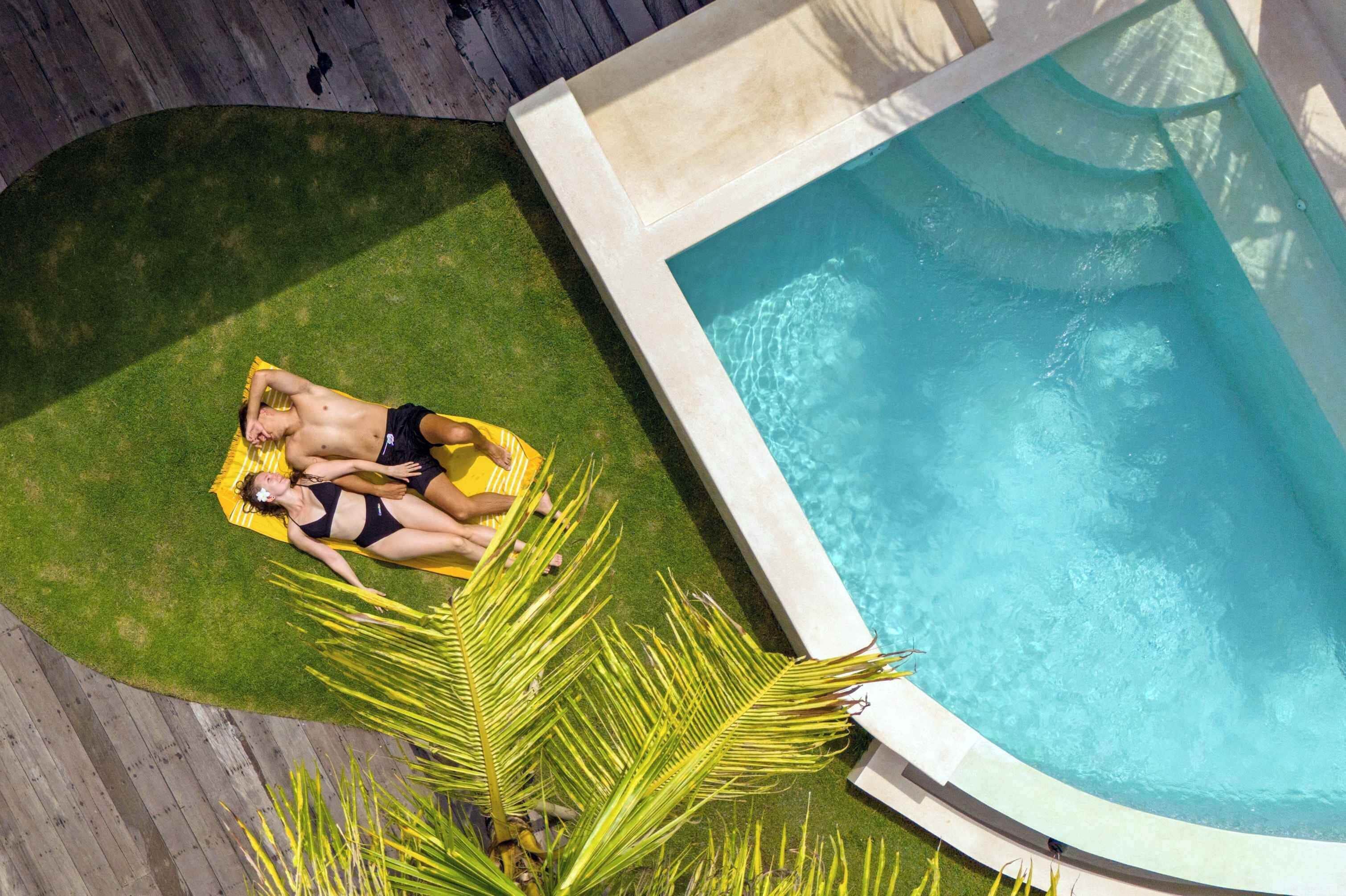 Ein Pool mit klarer, blauer Wasseroberfläche, eine Rasenfläche mit einer gelben Liege und eine Palme.
