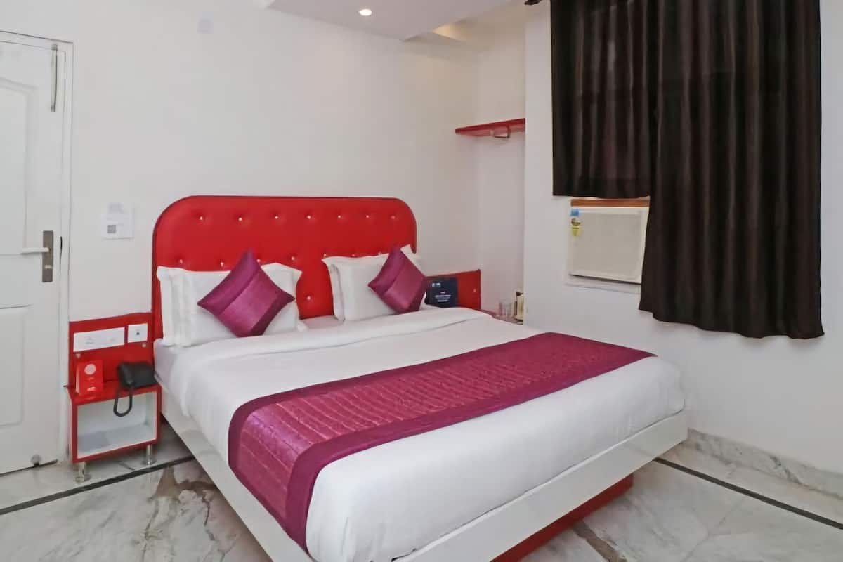 Quarto de hotel com cabeceira vermelha, roupa de cama branca, mesa de cabeceira vermelha e janela com cortinas escuras.