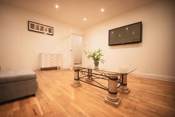 A spacious room with a wooden floor, a glass-top table with metal legs, a grey sofa, a white dresser, a potted plant, and a flat-screen TV mounted on the wall. - Stylish flat in Oxford Circus
(London)