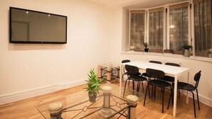 Un área de comedor con una mesa blanca y sillas negras, una mesa de centro de vidrio con una planta, y un televisor de pantalla plana grande montado en la pared.
