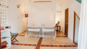 A room with a bed under a canopy, a small table with a chair, a shelf with items, and a door. - Beach front Mawimbi Studio in stunning garden (Coast)