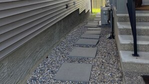 A narrow pathway with steps leading to a building with horizontal siding and a covered area with outdoor equipment. - Charming Oasis Tranquil Suite 8 min Airport, AC (Calgary)
