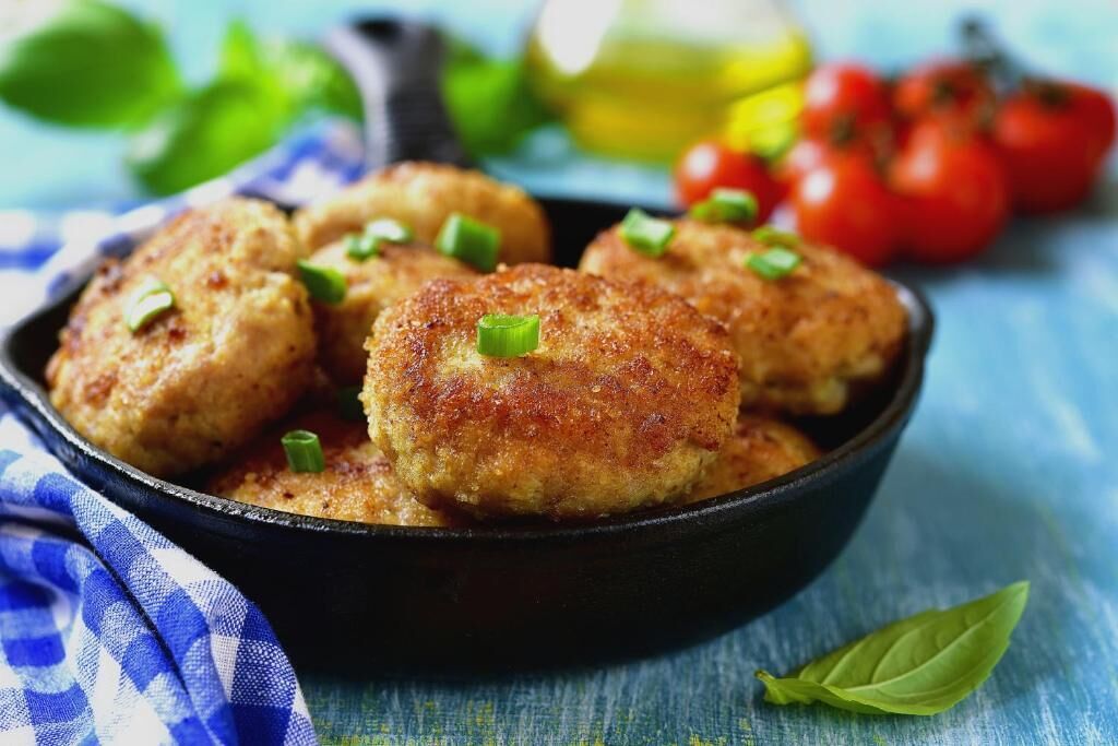 A skillet with crab cakes garnished with green onions, surrounded by tomatoes and basil leaves on a blue surface.