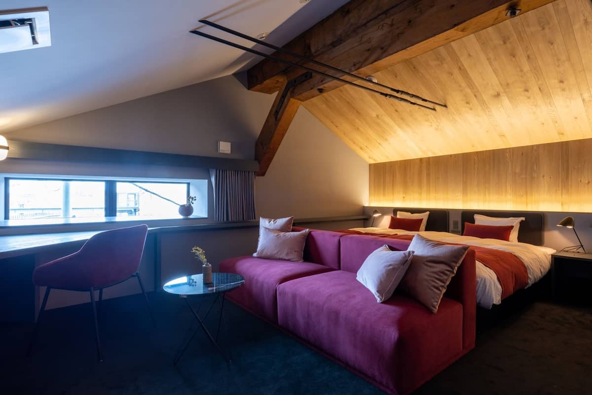 Ein modernes Schlafzimmer mit geneigter Decke, einem großen Bett mit roter Bettwäsche, einem roten Sofa, einem kleinen runden Tisch und einem Fenster mit Vorhängen.
