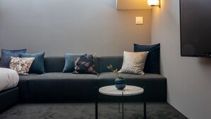 A modern living room with a dark grey sofa, multiple cushions, a small round table with a vase, and a flat-screen TV mounted on the wall. - NIPPONIA HOTEL Hakodate Port Town (Hakodate)