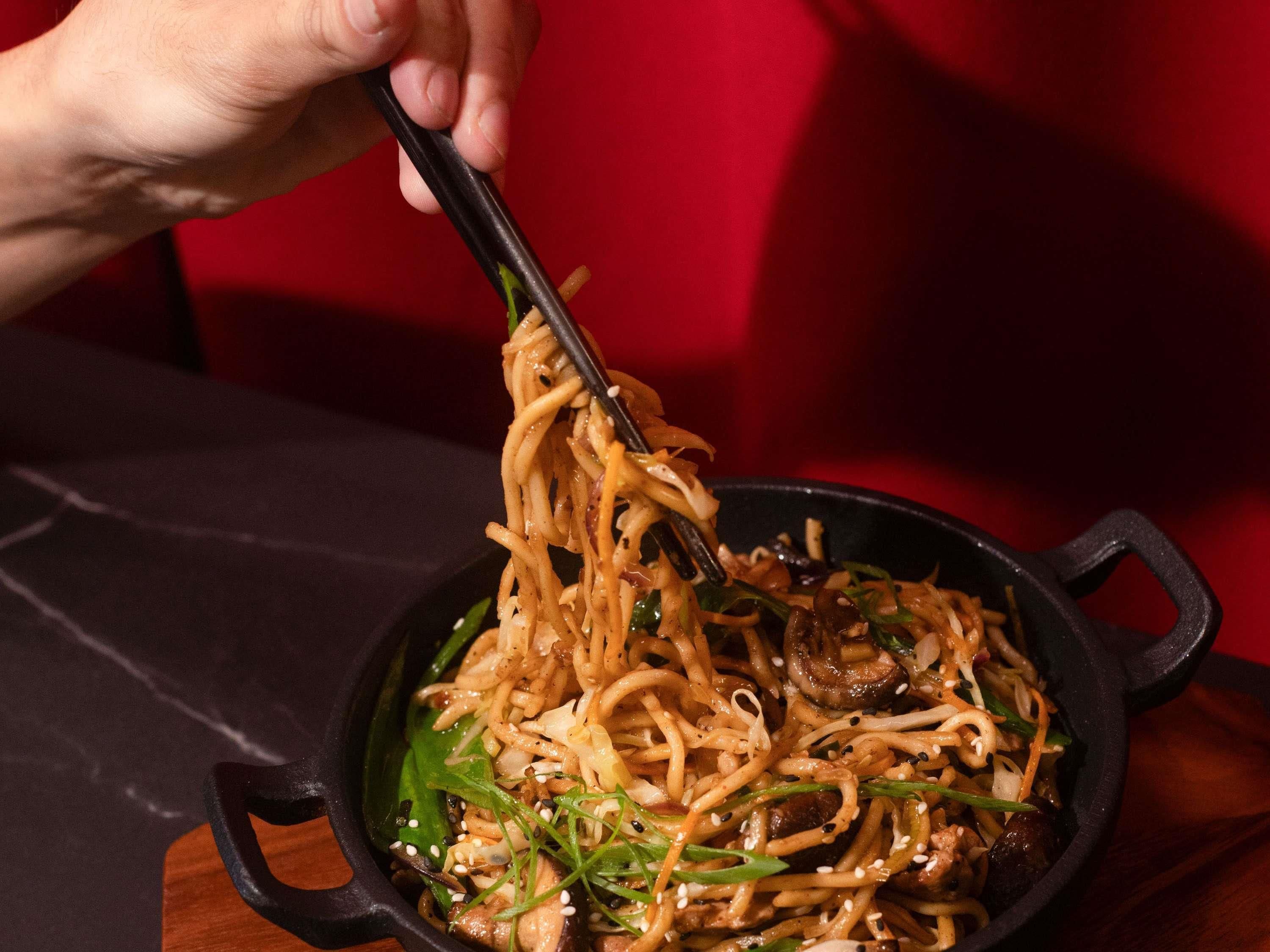 A person holding chopsticks over a bowl of noodles with vegetables and mushrooms.