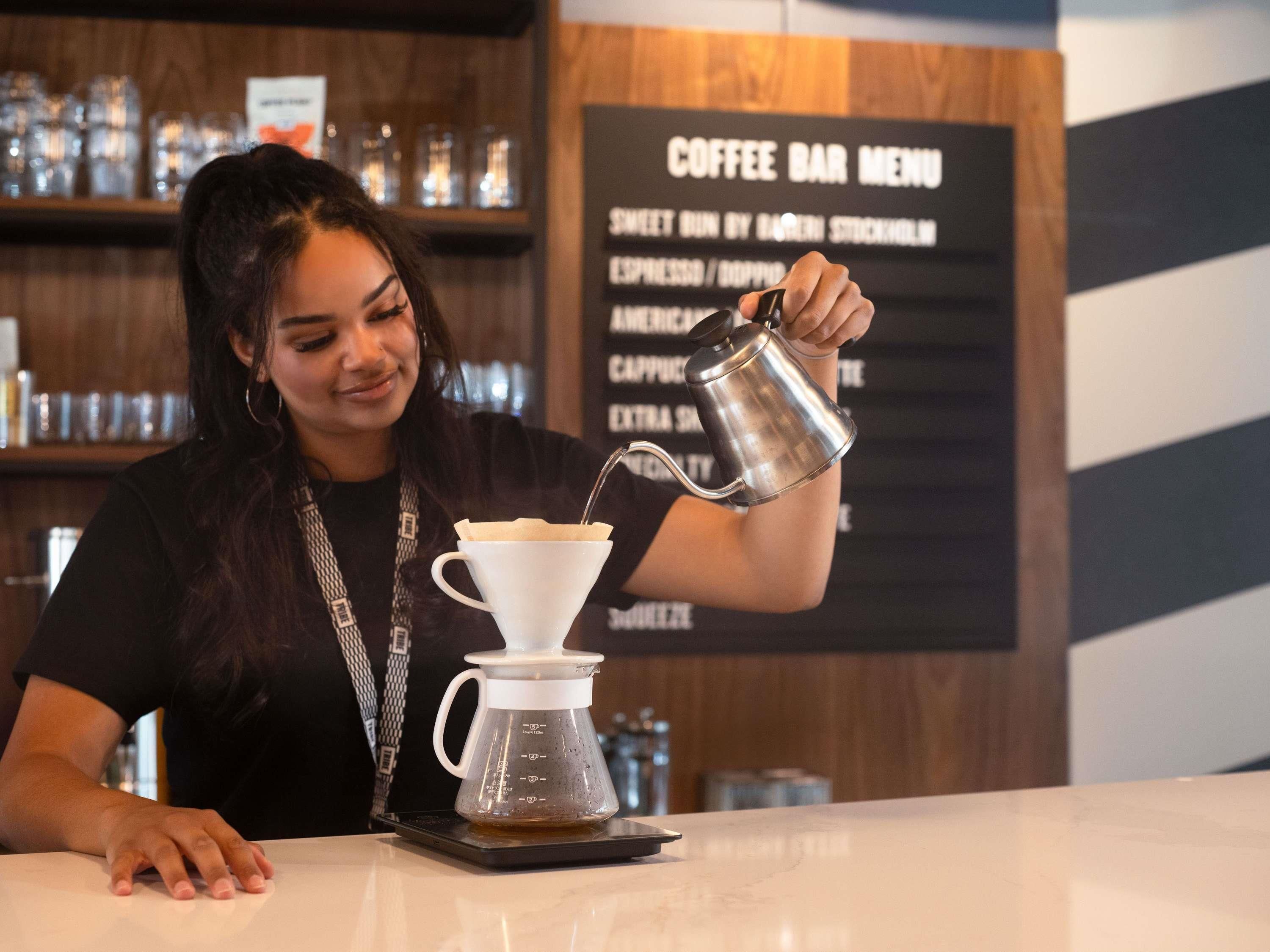 Una persona sirve café en una taza en una cafetería, con un menú de fondo.