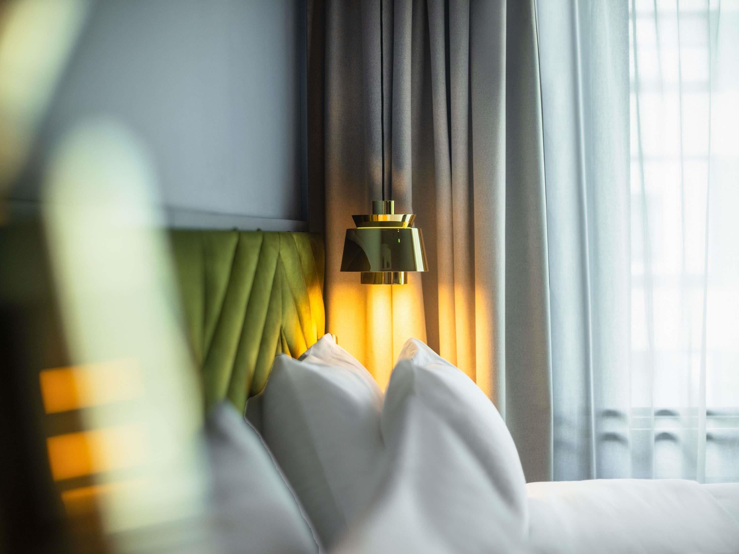 A hotel room with a bed, a green headboard, a wall-mounted lamp, and a window with curtains.