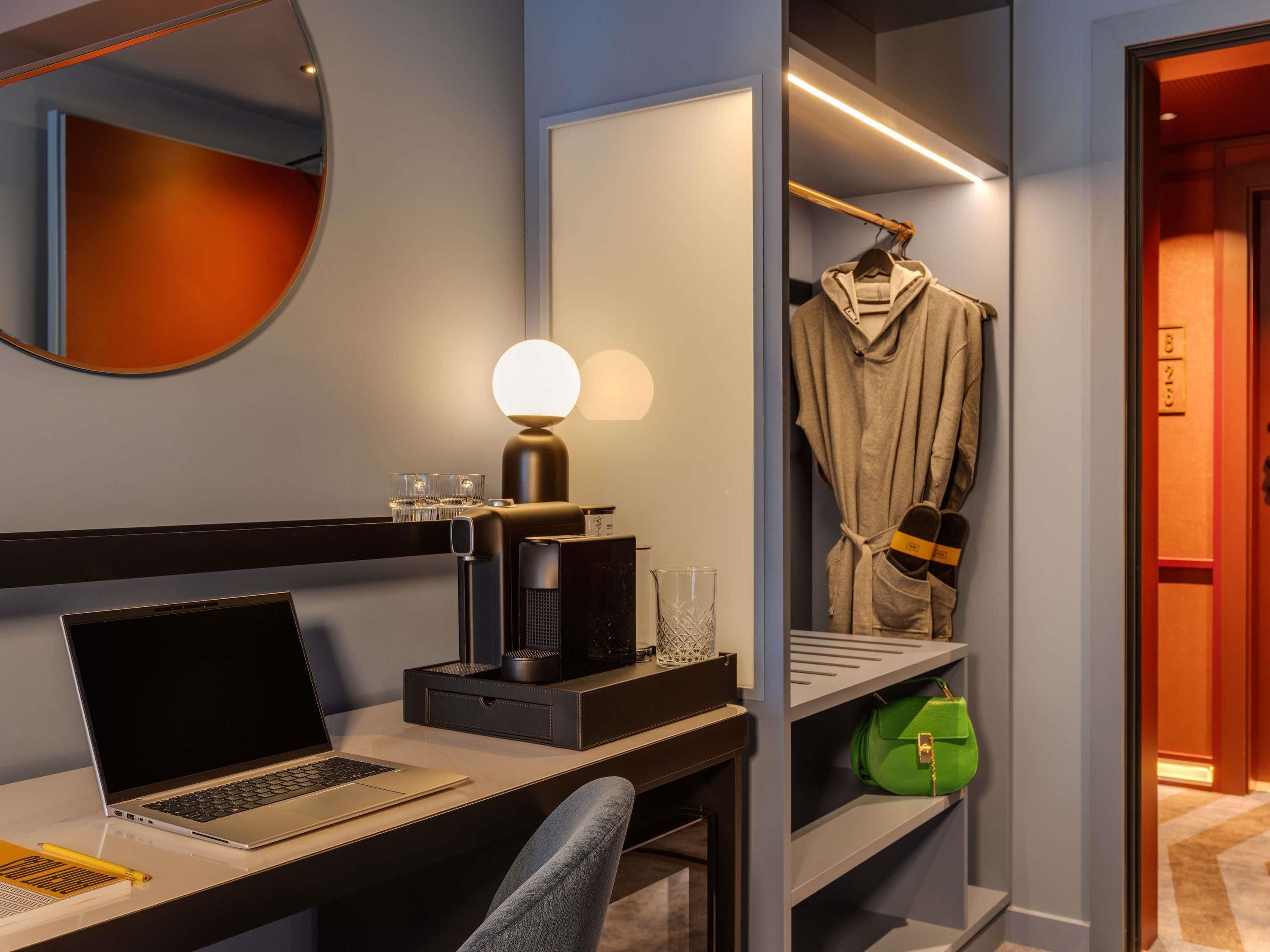 A hotel room with a desk, laptop, lamp, and a robe hanging on a rack.