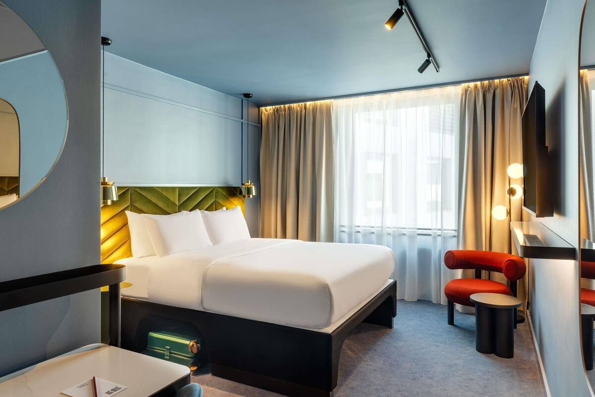 A hotel room with a bed, a desk with a chair, a small table, a mirror, a red chair, and a window with curtains.