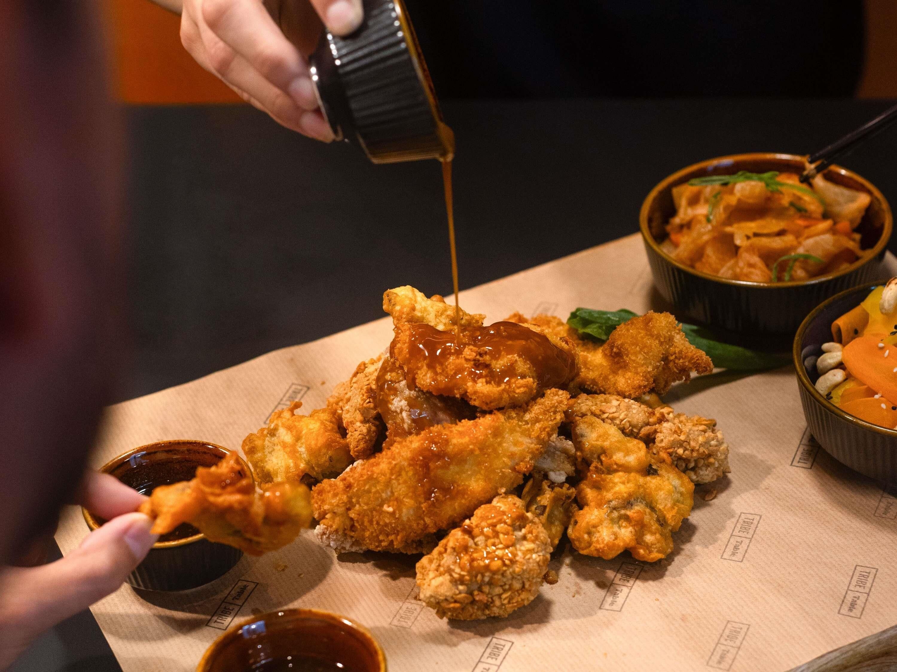 Eine Person gießt Soße über gebratene Chicken Wings auf einem Tisch.