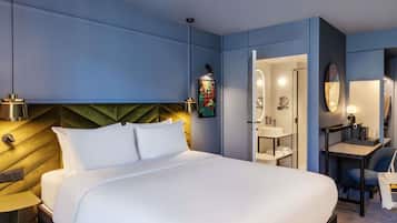 A modern hotel room with a large bed, bedside tables, a desk with a chair, a bathroom with a mirror and sink, and a painting on the wall.