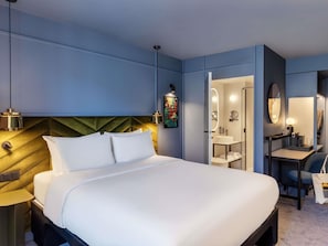 Ett modernt hotellrum med en stor säng, sängbord, ett skrivbord med en stol, en spegel och ett badrum med handfat och spegel.