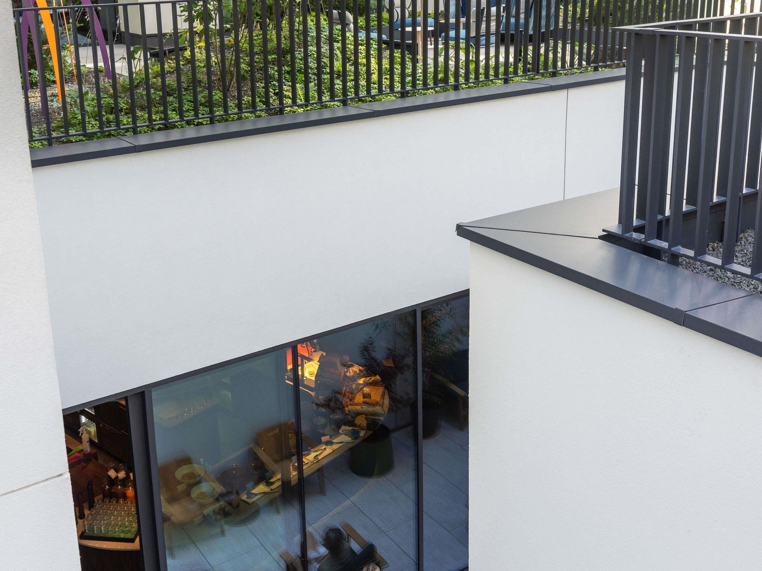 En modern byggnad med en glasfasad, en balkong med ett metallräcke och en trädgårdsyta som syns genom räcket.