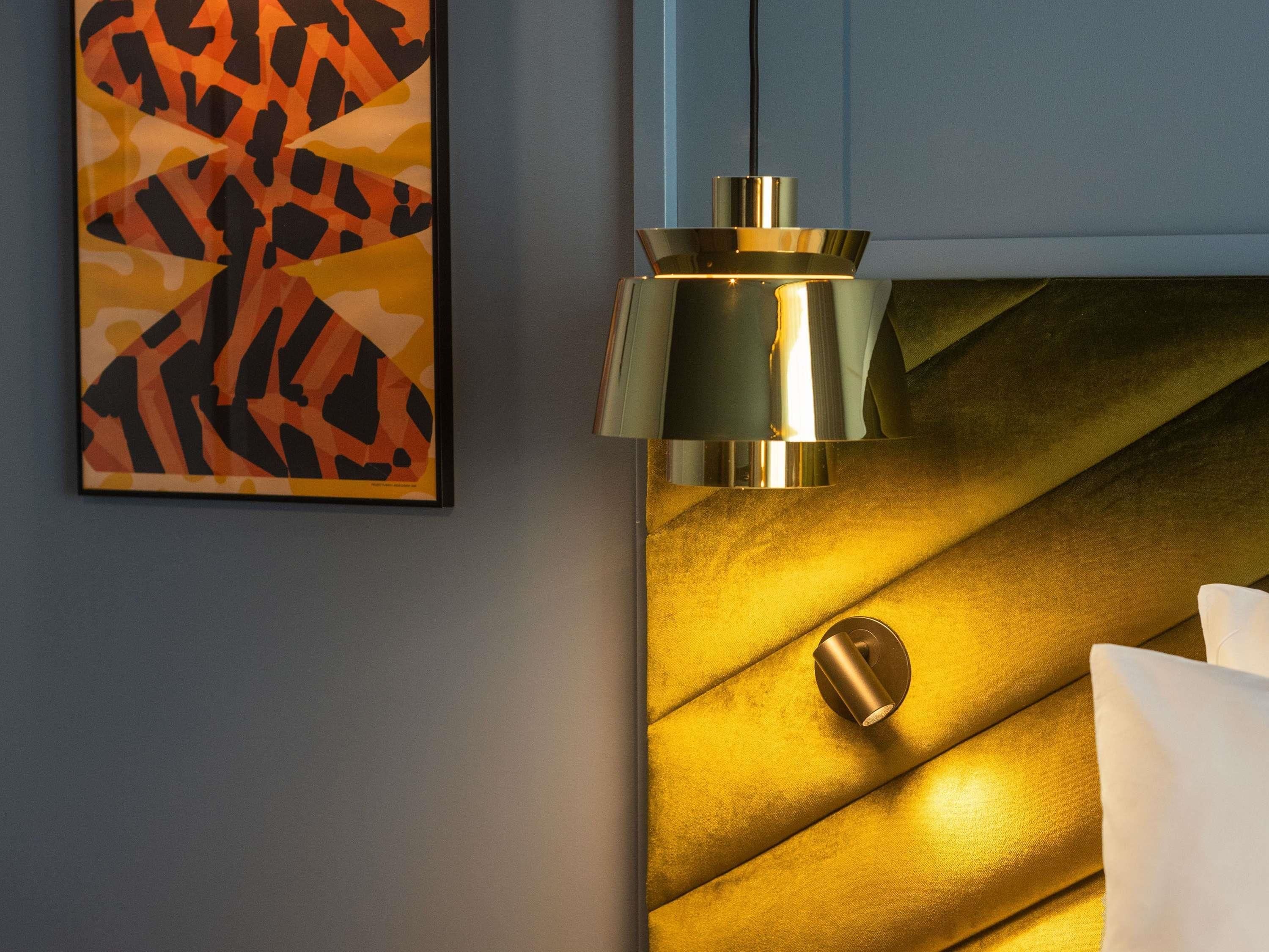 A room with a modern design, featuring a geometric patterned artwork on the wall, a metallic light fixture, and a textured upholstered headboard.