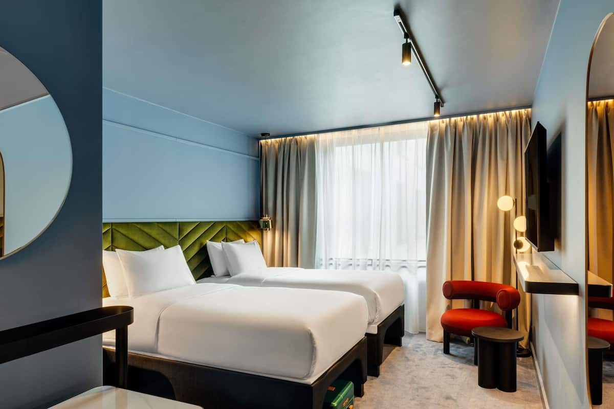 A hotel room with two beds, a desk, a chair, a mirror, and a window with curtains.