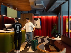Un moderno vestíbulo de hotel con una mesa de recepción, una zona de estar con un sofá, una maleta verde y un ordenador portátil sobre el mostrador.