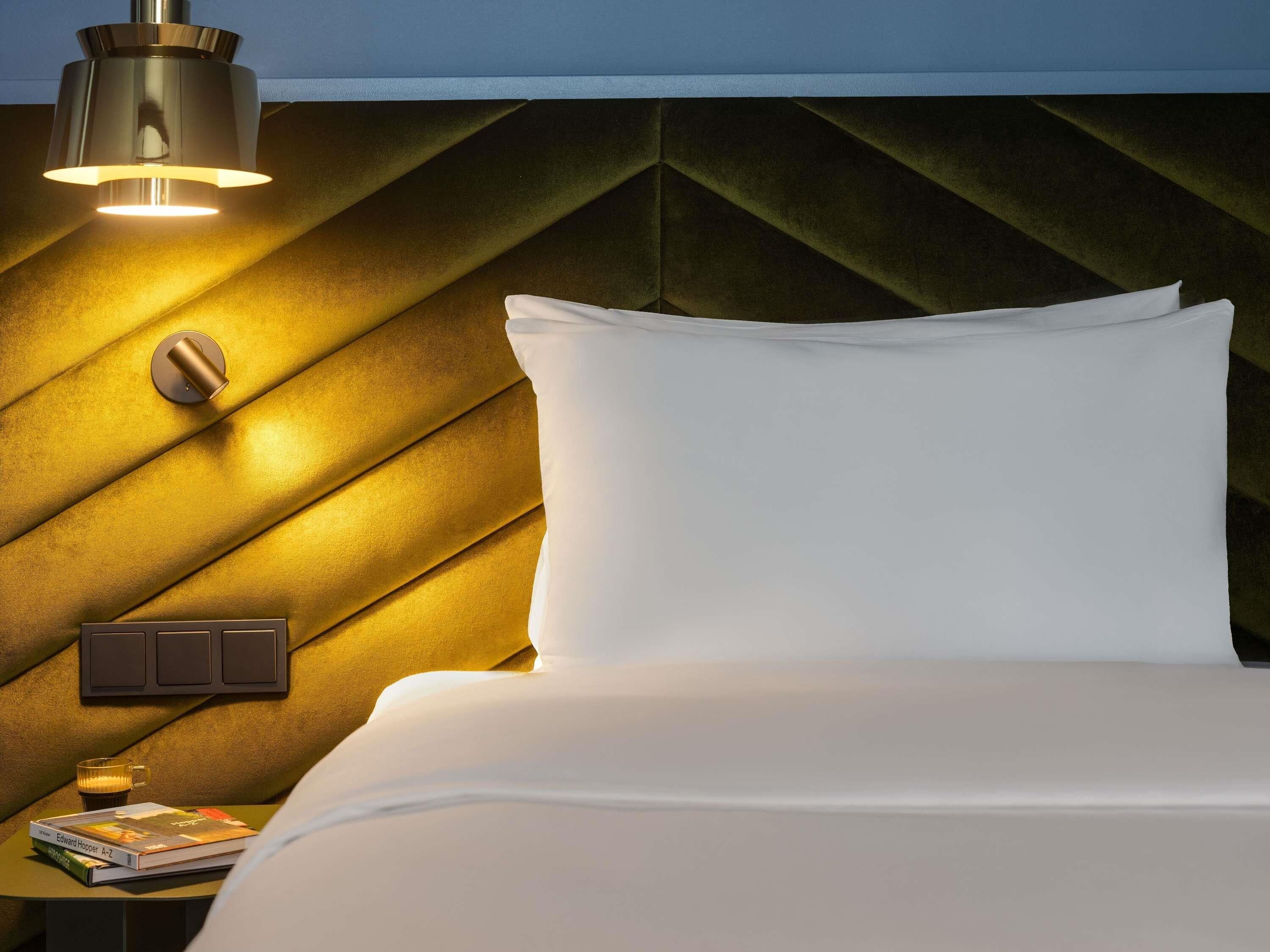 Una cama con almohadas blancas, una mesita de noche con un libro y una taza, y una lámpara fijada a la pared.