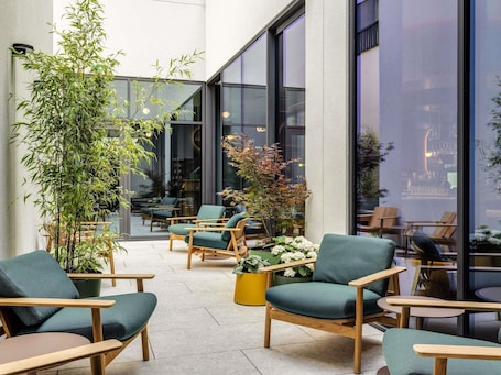 A modern indoor seating area with wooden chairs and green cushions, surrounded by large windows and indoor plants.. TRIBE Krakow Old Town