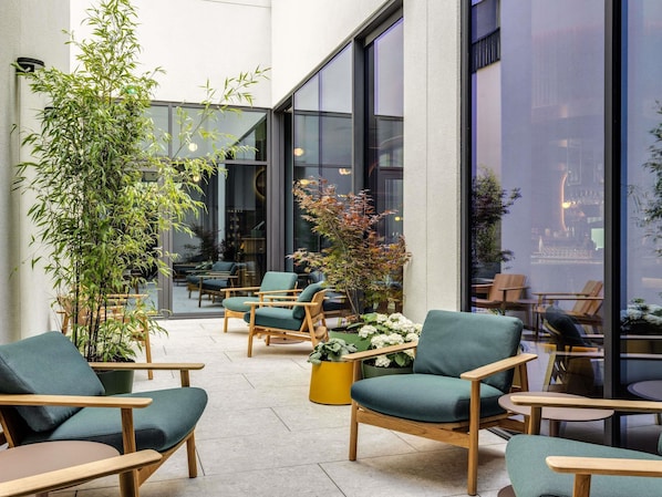 Un área de estar interior moderna con sillas de madera y cojines verdes, rodeada de grandes ventanales y plantas de interior.