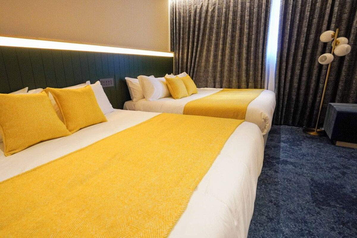 Habitación de hotel con dos camas, cada una con colcha y almohadas amarillas, cabecero oscuro, alfombra azul y una lámpara de pie.