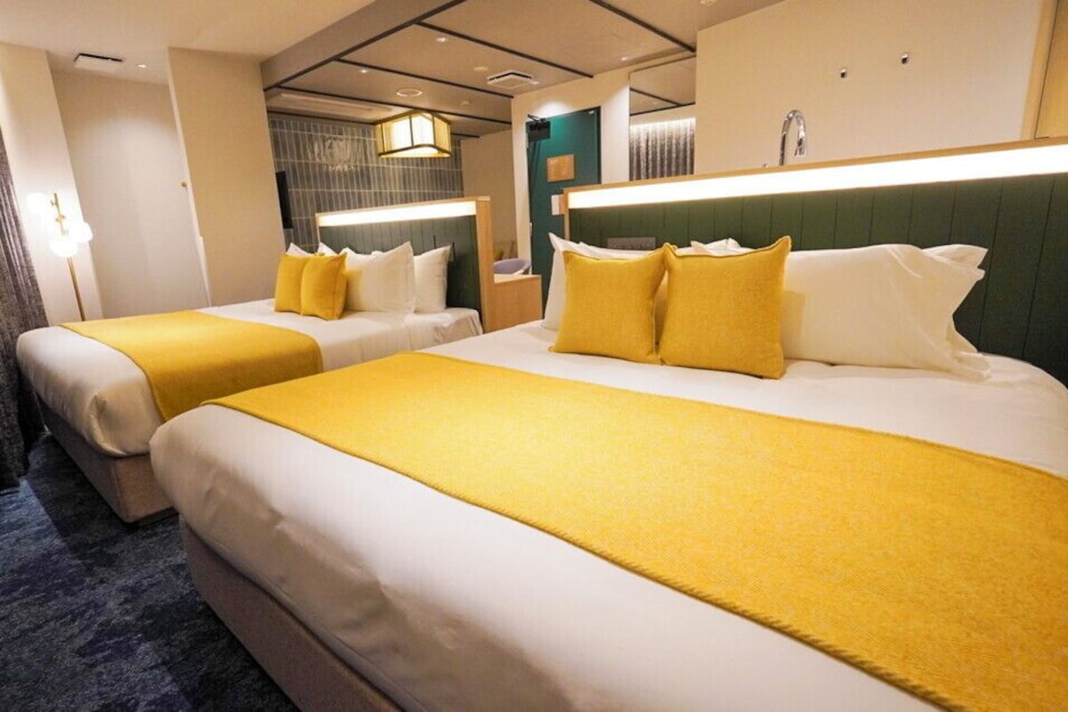 Habitación de hotel con dos camas, cada una con ropa blanca y detalles amarillos, un cabecero moderno, un ventanal grande y un baño visible con lavabo y espejo.