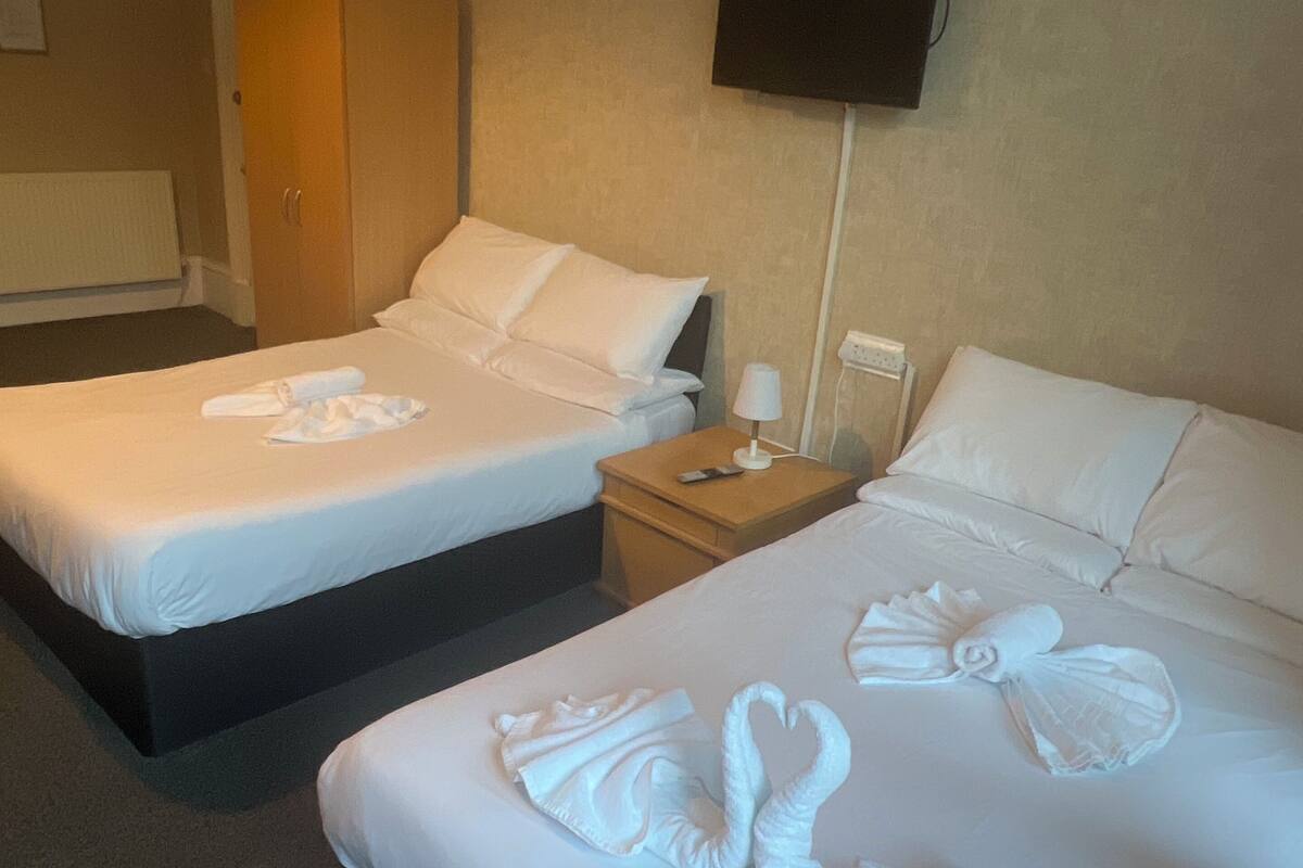 Two beds with white linens and towels arranged in a swan shape. There is a wooden nightstand between the beds, and a flat-screen TV mounted on the wall above the beds.
