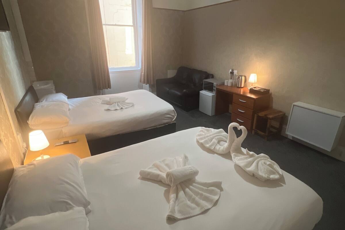 A hotel room with two beds, a desk with a lamp, a microwave, a small table with a lamp, and a chair.