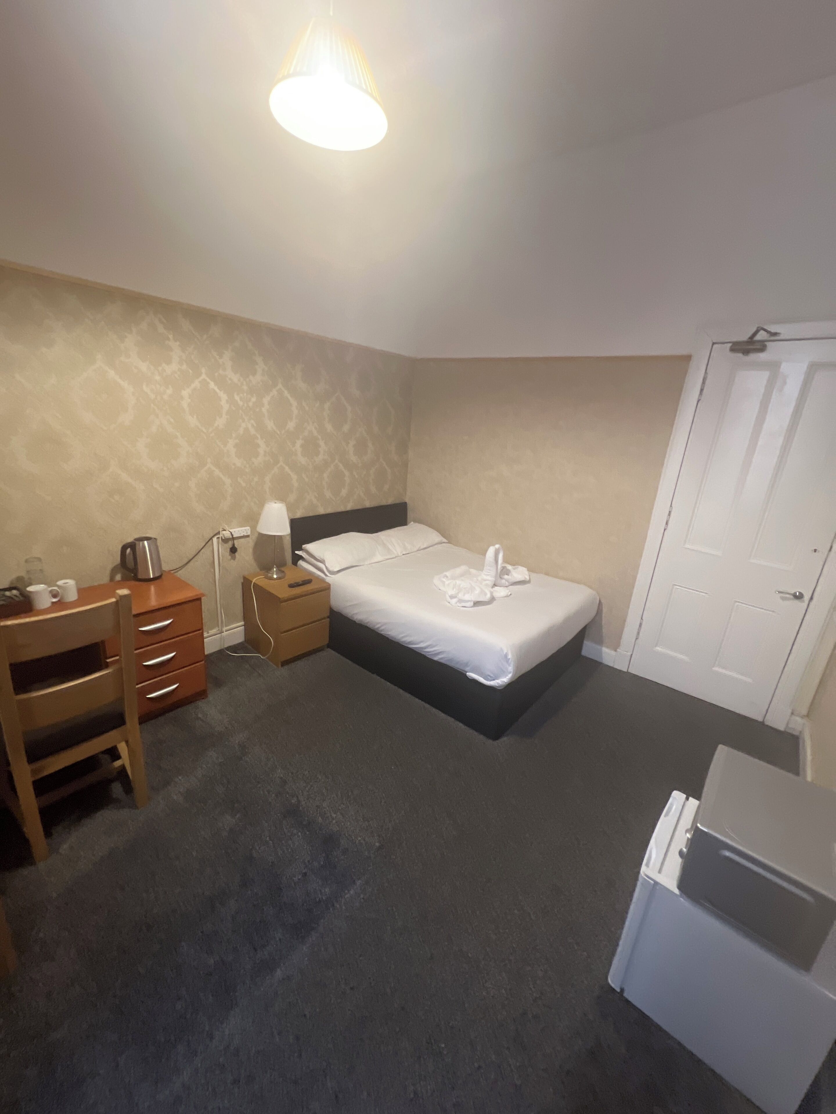 A hotel room with a bed, a desk, a chair, a nightstand, a lamp, a coffee maker, and a wardrobe.