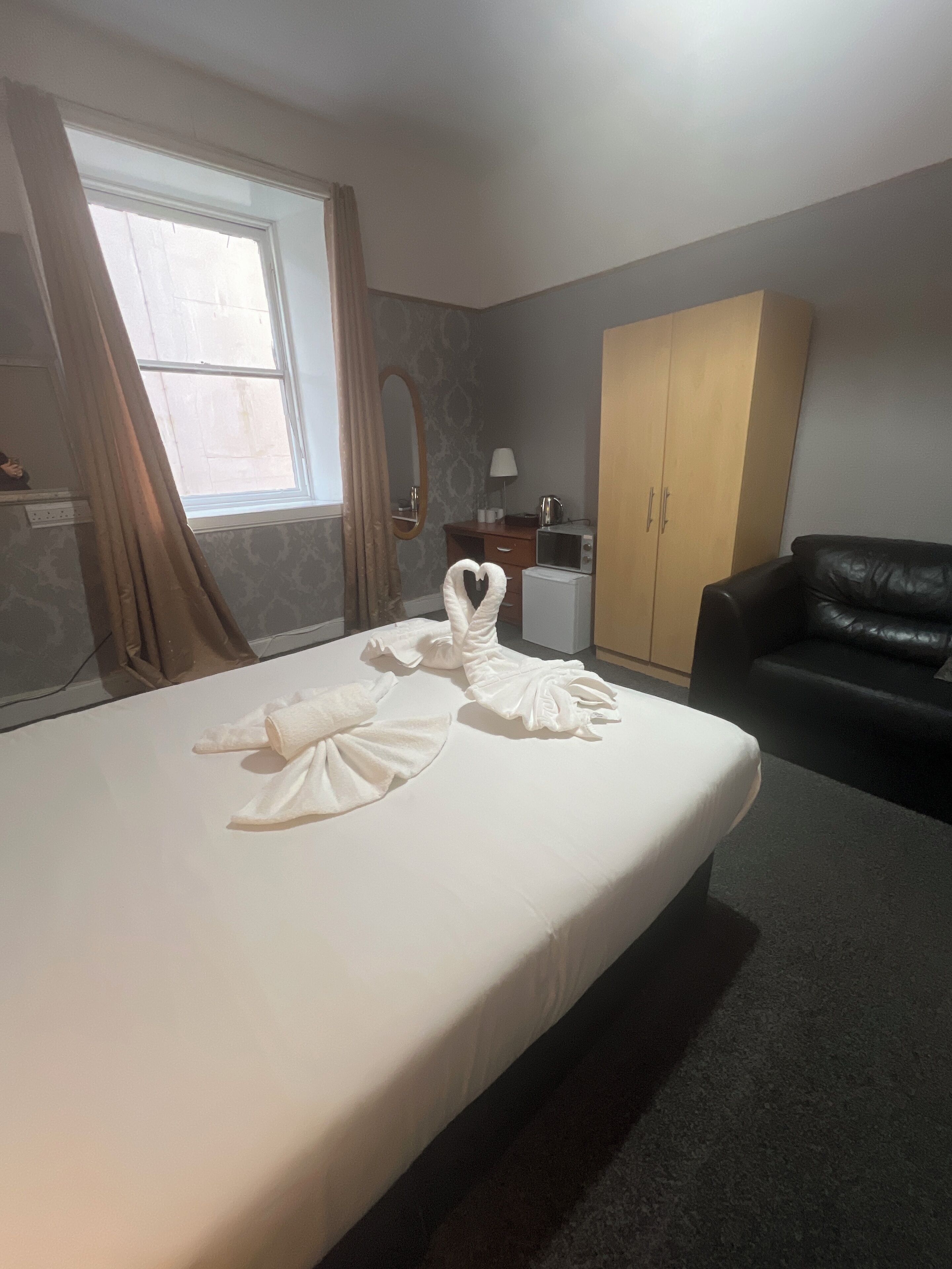 A hotel room with a neatly made bed, a black leather sofa, a wooden wardrobe, a small desk with a lamp, and a window with curtains.