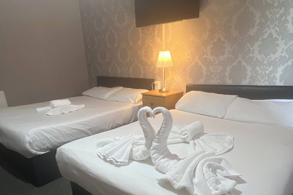 Two beds with white linens and towels arranged in a swan shape. There is a nightstand with a lamp between the beds.