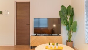 A modern living room with a flat-screen TV on a wooden cabinet, a large potted plant, and a small table with decorative items. - Travel Time Sumiyoshi Suzaku (Osaka)