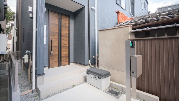 A modern residential building with a wooden door, a small step leading to the entrance, and a grey utility box near the door. - Travel Time Sumiyoshi Suzaku (Osaka)