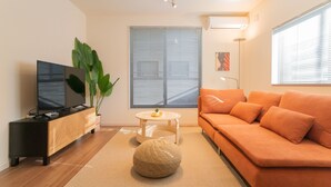 A modern living room with a sofa, a TV on a wooden stand, a round ottoman, a small table with a plant, and a large window with blinds. - Travel Time Sumiyoshi Suzaku (Osaka)