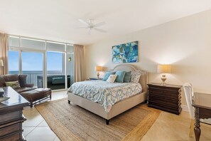 Ein Schlafzimmer mit einem großen Bett, einem Deckenventilator, einem Nachttisch mit Lampe, einer Kommode, einem Sessel und Blick ins Freie durch eine Schiebetür.