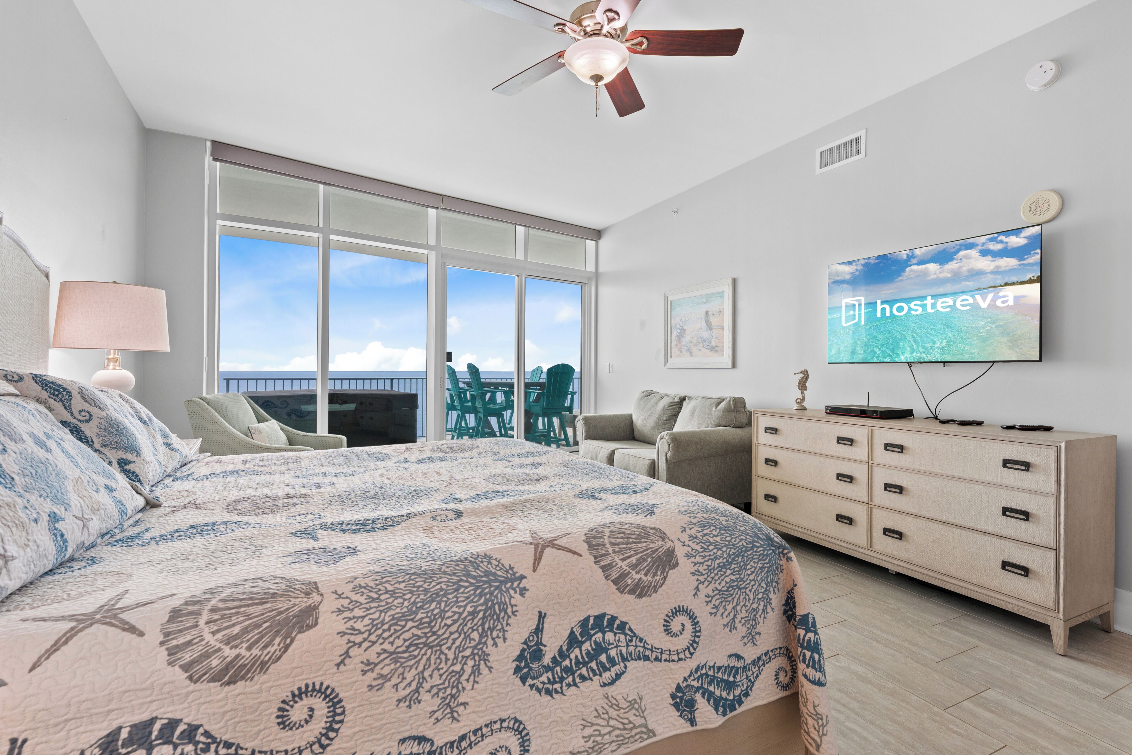 A bedroom with a large bed, a ceiling fan, a TV, a dresser, and a view of the ocean.