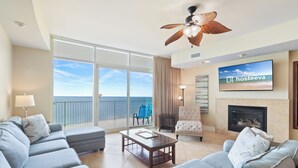 A spacious living room with a large sofa, a coffee table, and a fireplace. There is a television mounted on the wall and a ceiling fan above. - Turquoise Place Condos (Orange Beach)