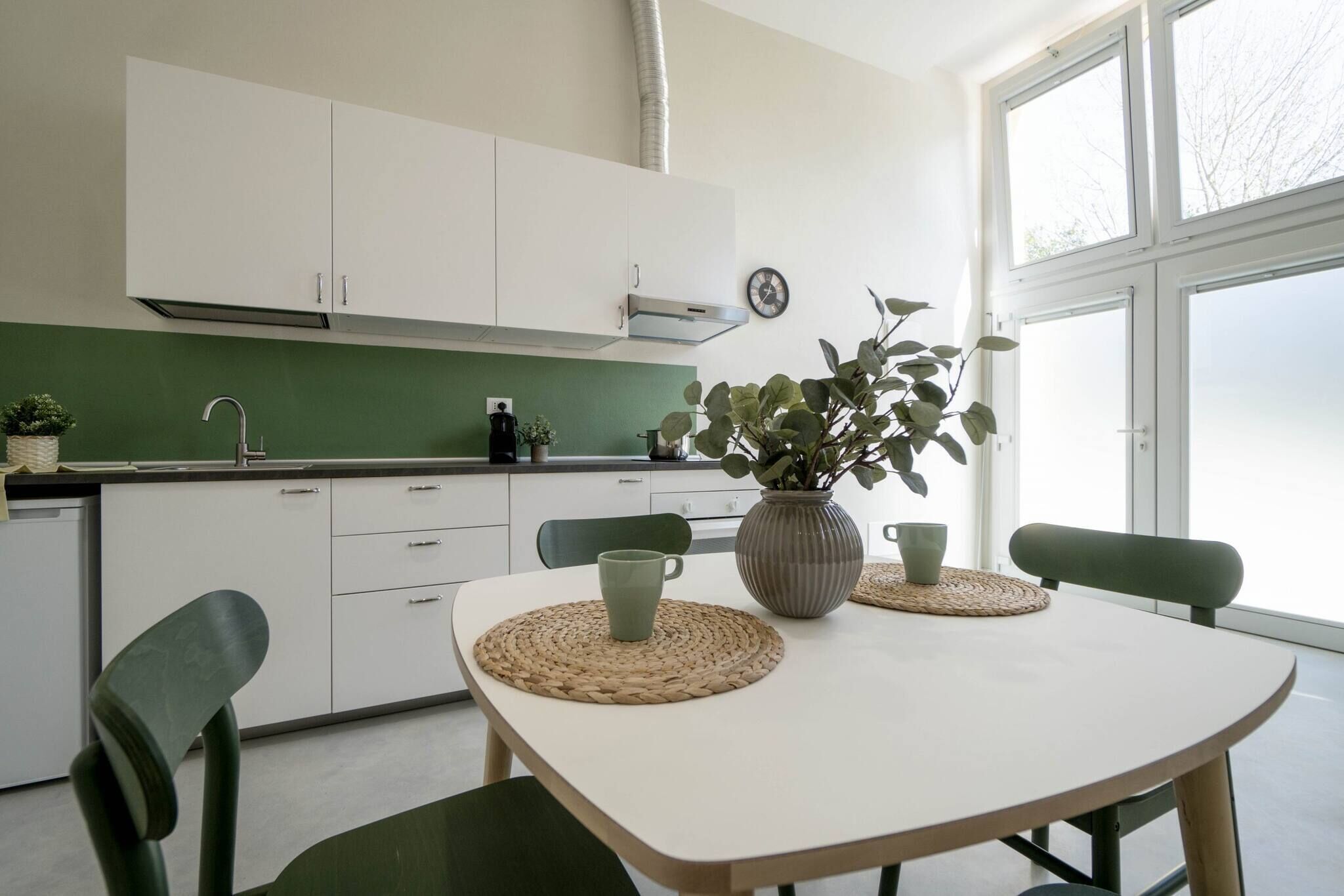 A modern kitchen with white cabinets, a green backsplash, and a dining table with chairs. There is a clock on the wall and a window providing natural light.