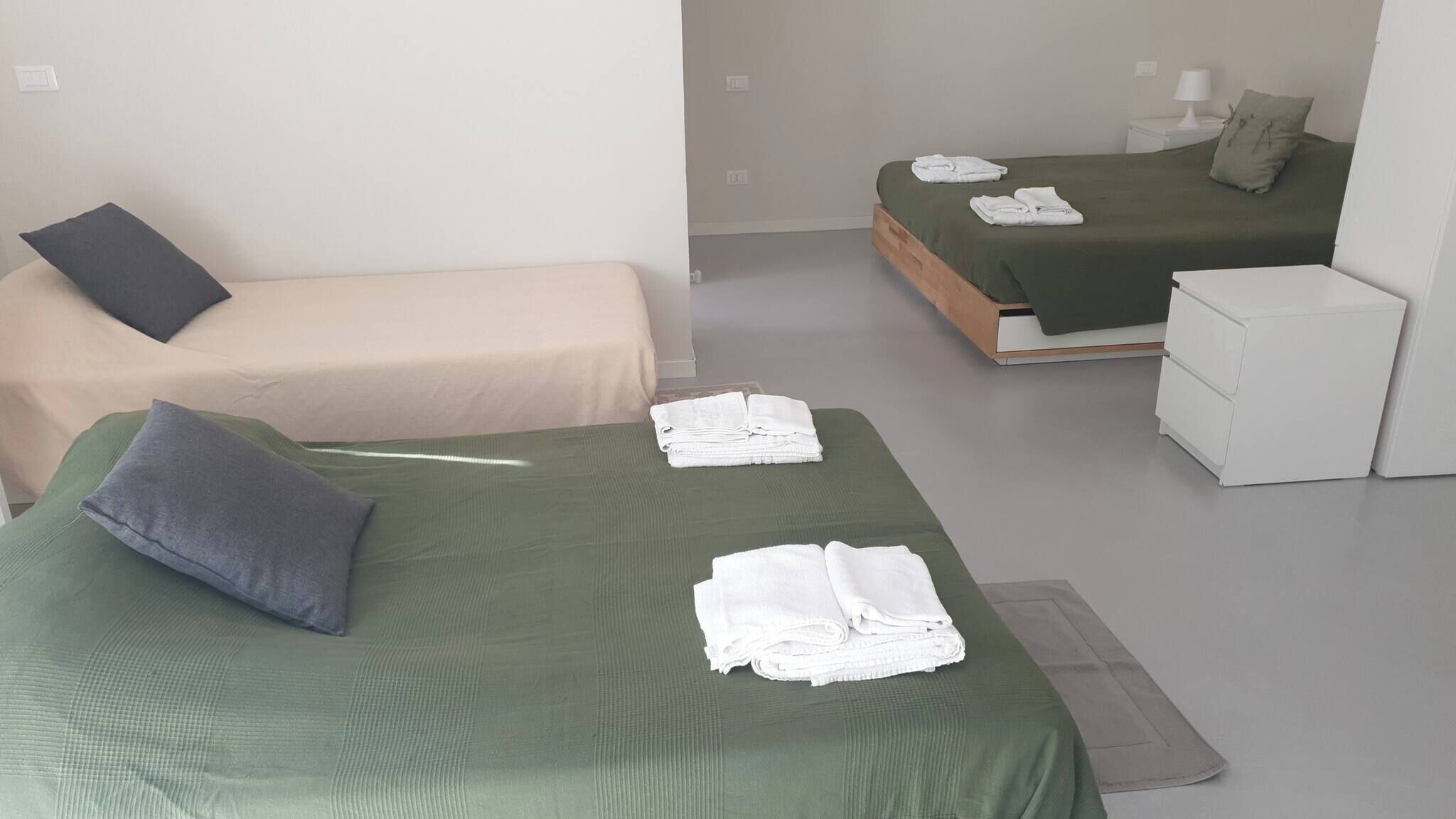 A hotel room with two beds, one with a green bedspread and the other with a beige bedspread. There are towels on the beds and a small white cabinet.